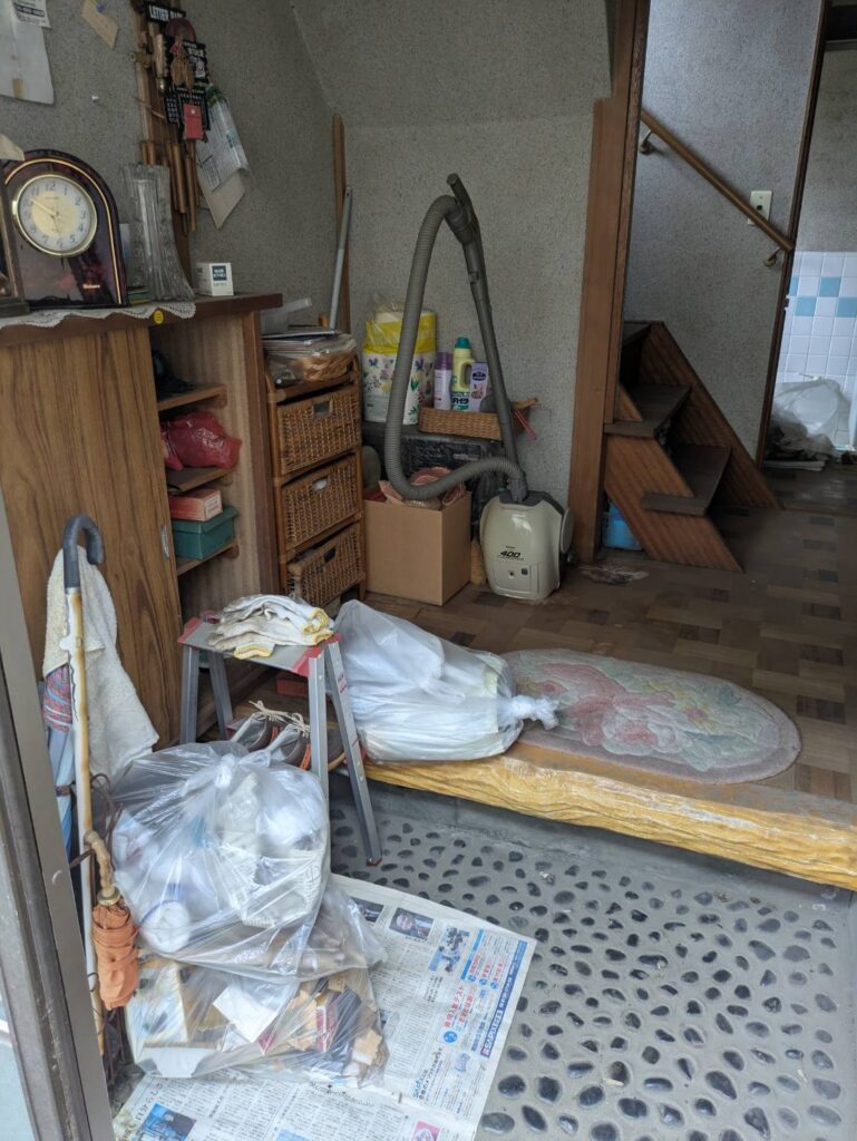 作業前の玄関。上がり口に袋に入った日用品や掃除機、棚が残っている