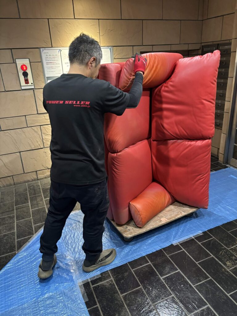 床にブルーシートを敷いたエントランスで、赤いソファを台車に載せて運ぶ当社スタッフ